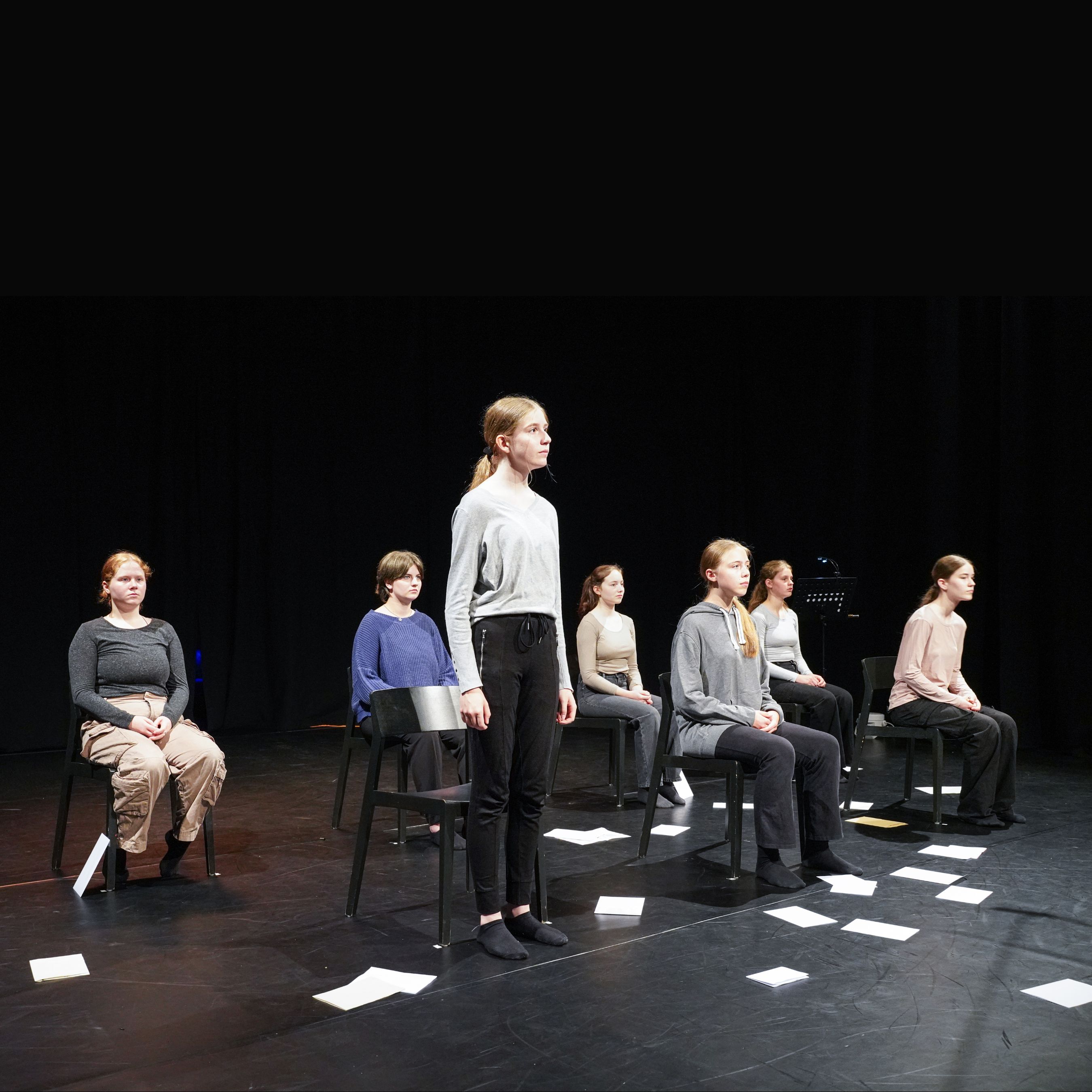Bühnenbild mit sieben schwarzen Stühlen in zwei Reihen. Eine Frau steht. Die anderen sechs Frauen sitzen.